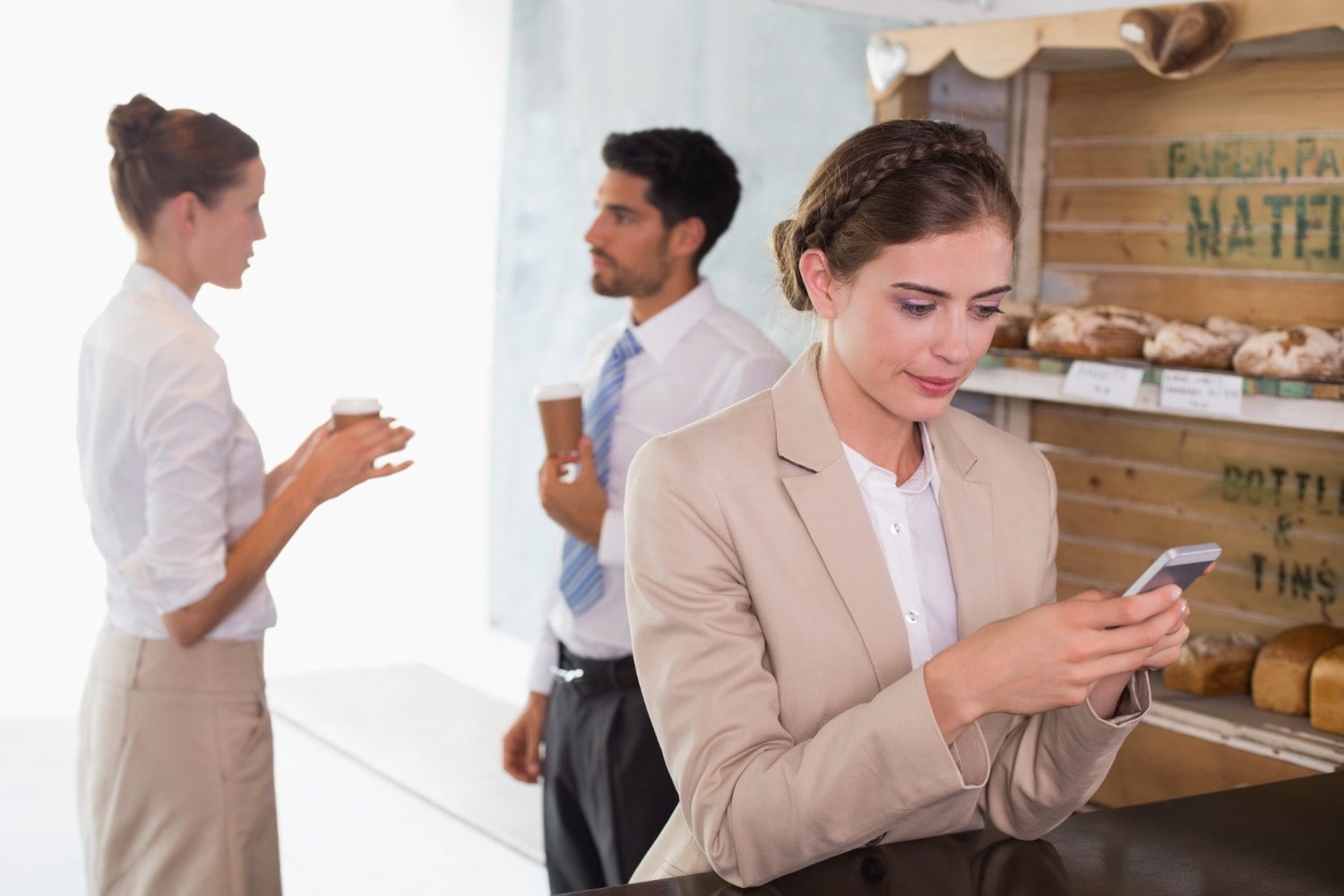 A businesswoman in a beige blazer checks her phone while standing near a bakery counter, showcasing the power of seamless communication tools. In the background, two colleagues converse over coffee, highlighting unified communications for effective professional interactions.