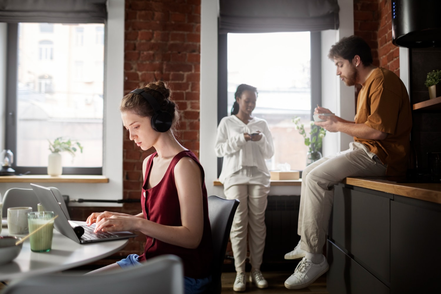 A remote work setting features a woman in headphones focusing on her laptop, while two colleagues chat nearby in a cozy, modern kitchen. This scene represents the flexibility of business telecom solutions and seamless communication tools in supporting unified communications and team productivity across different environments.