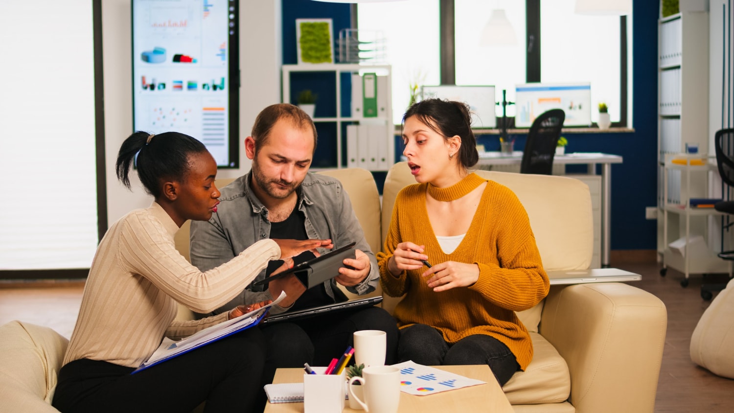 A collaborative team meeting takes place in a casual office setting, with three individuals actively discussing a project using a tablet. Their use of technology underscores the importance of unified communications and business telecom solutions for enhanced team productivity.