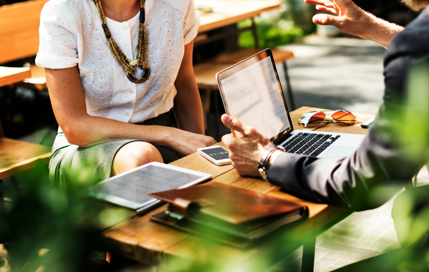 Two professionals in an outdoor café setting discuss data on a laptop, surrounded by a tablet and smartphone, symbolizing scalable IT infrastructure and the need for fast business internet in mobile and collaborative work environments.