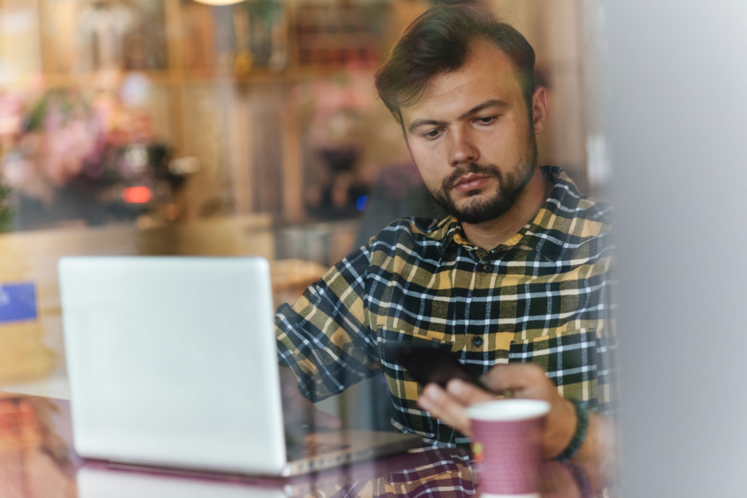 A man in a plaid shirt sits at a café using a smartphone and laptop, conveying the role of reliable internet solutions and business internet access for remote professionals managing tasks on the go.