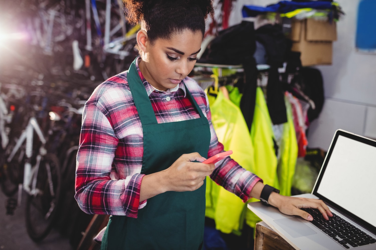 A woman in a green apron at a retail store uses a smartphone while working on a laptop, illustrating how scalable IT infrastructure and fast business internet enable seamless point-of-sale and inventory operations.