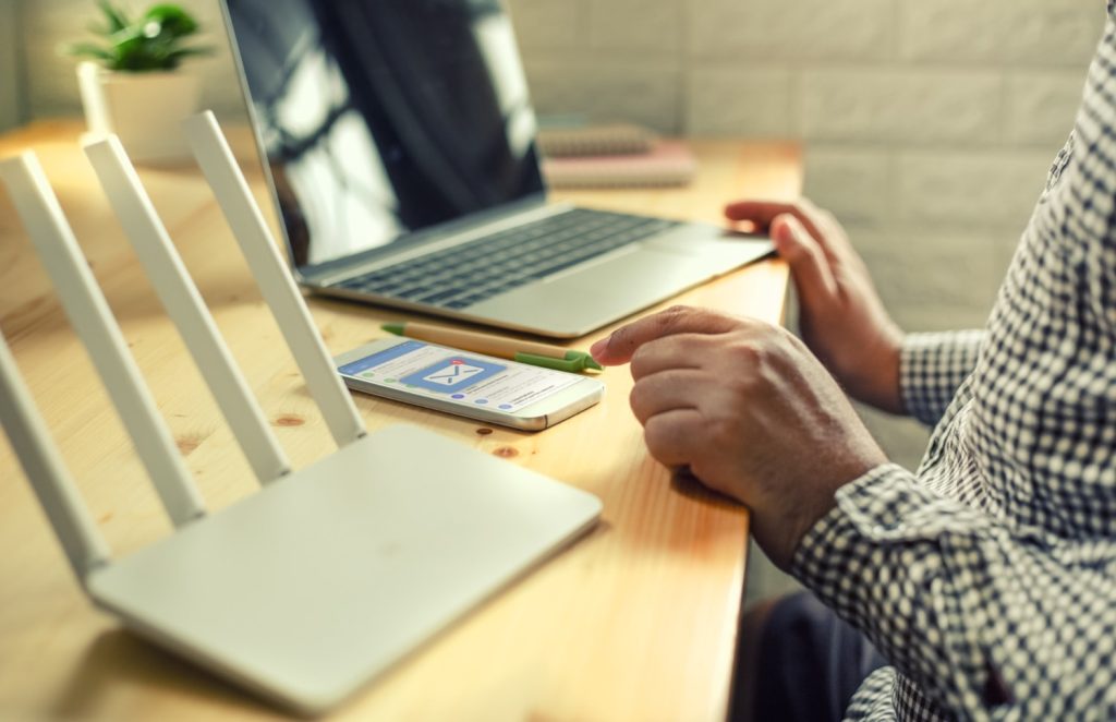Current image: A person at a wooden desk uses a smartphone displaying email notifications, with a laptop and white Wi-Fi router nearby, representing the importance of fast business internet and reliable internet solutions for remote work efficiency.