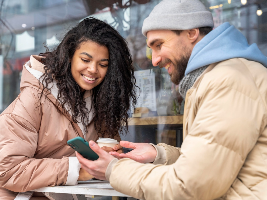 A smiling woman in a pink winter coat and a man in a beige jacket and gray beanie sit at an outdoor café table, looking at a smartphone together. They appear to be enjoying a moment of casual internal business messaging using mobile messaging solutions.